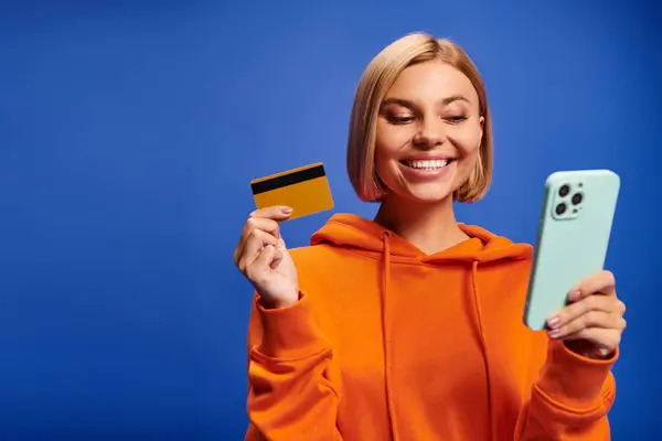 Atractiva mujer alegre con el pelo rubio en sudadera naranja vibrante posando con tarjeta de crédito y teléfono - foto de stock