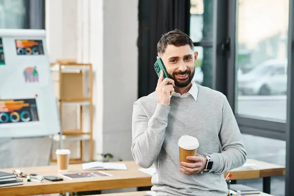 Un hombre de negocios en una oficina corporativa multitarea hablando en un teléfono celular mientras sostiene una taza de café. - foto de stock