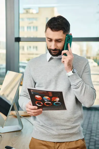 Um homem em um ambiente de negócios detém gráficos em uma mão e fala em um telefone celular na outra, multitarefa para o trabalho. — Fotografia de Stock