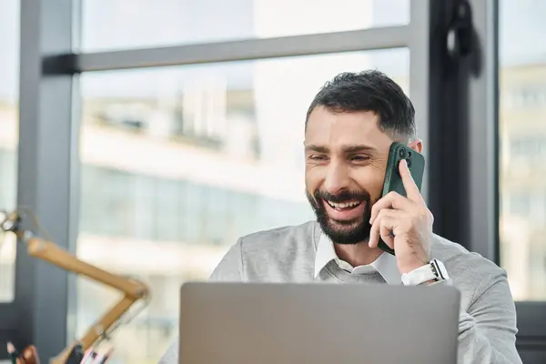 Un hombre se sienta frente a un portátil, hablando por un teléfono celular, inmerso en su trabajo en una oficina bulliciosa. - foto de stock