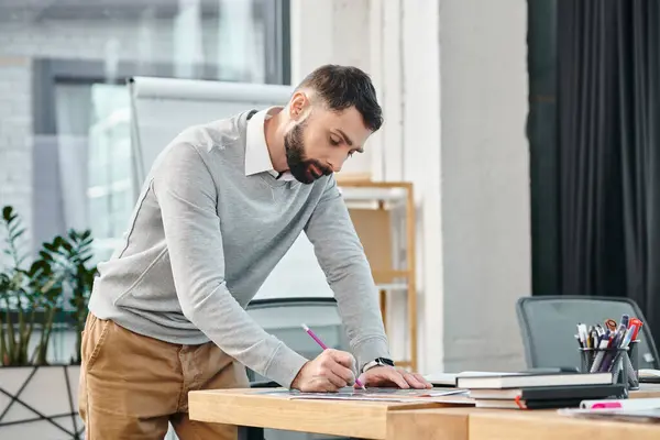 Un hombre parado en un escritorio, enfocado, escribiendo en un pedazo de papel con determinación mientras trabaja en un proyecto en una oficina corporativa. - foto de stock