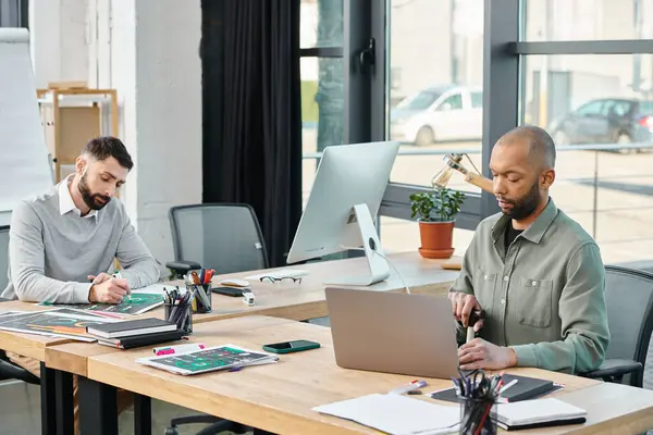 Deux hommes d'affaires sont assis à une table, immergés dans leurs ordinateurs portables, faisant du remue-méninges et collaborant à un projet dans un cadre de bureau moderne, inclusif — Photo de stock