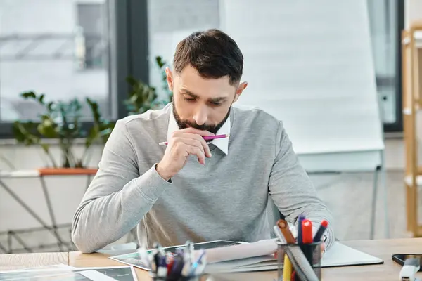 A man sits at a desk, engrossed in a piece of paper, deep in thought, surrounded by the hustle of a corporate office. — Photo de stock