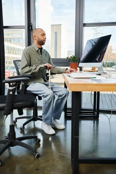 Hombre afroamericano discapacitado con miastenia gravis en traje corporativo se sienta enfocado en un escritorio, escribiendo en una pantalla de computadora entre los suministros de oficina. - foto de stock