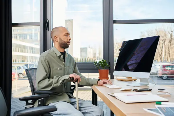 Hombre afroamericano discapacitado con miastenia gravis se sienta en un escritorio absorto en su trabajo, frente a una pantalla de computadora en un entorno de oficina típico de la cultura corporativa. - foto de stock