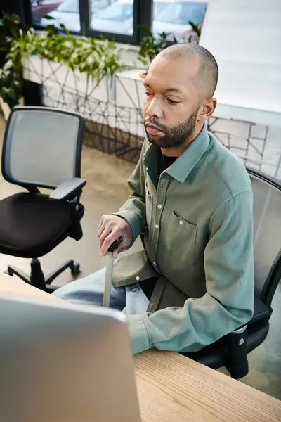 Behinderter afrikanisch-amerikanischer Mann mit Myasthenia gravis vor einem Laptop, konzentriert auf die Arbeit in einem geschäftigen Büroumfeld voller Unternehmenskultur. — Stockfoto
