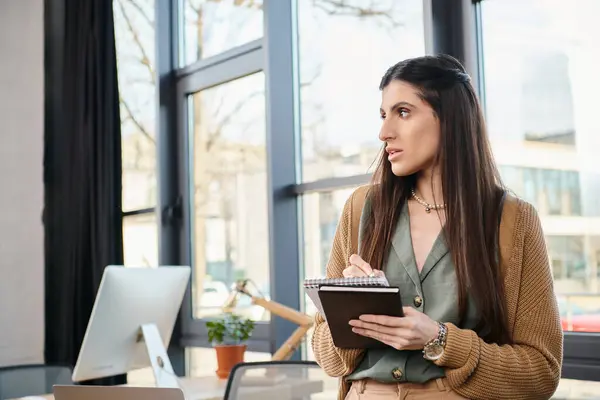 Женщина стоит перед компьютером, держа планшет, погруженная в свою работу в шумную офисную атмосферу. — стоковое фото