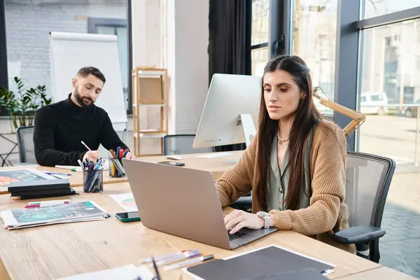 Мужчина и женщина, сидящие за столом, глубоко в работе на ноутбуках, воплощающие суть командной работы в корпоративной обстановке. — стоковое фото