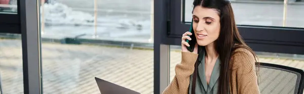 Una mujer que se dedica a una conversación en un teléfono celular mientras está sentada en una mesa en un entorno empresarial. - foto de stock