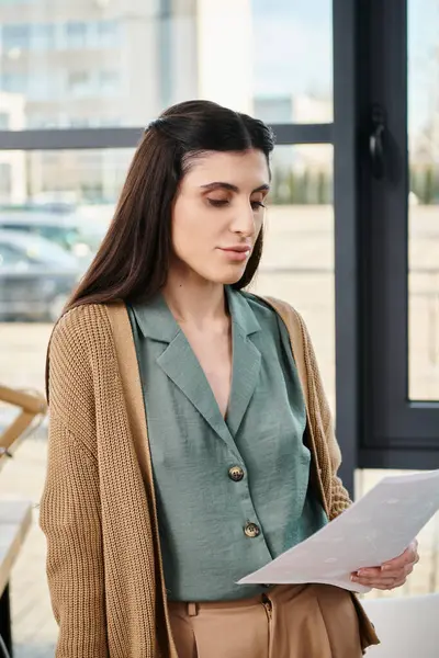 A woman standing by a window, holding a paper, reflecting on business ideas in a corporate office setting. — Stock Photo