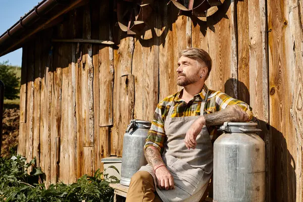 Atractivo granjero en traje casual con tatuajes sosteniendo batido de leche y mirando hacia otro lado al lado de la casa - foto de stock