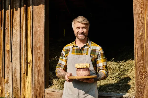 Alegre granjero guapo con tatuajes sosteniendo queso casero en sus manos y sonriendo a la cámara - foto de stock
