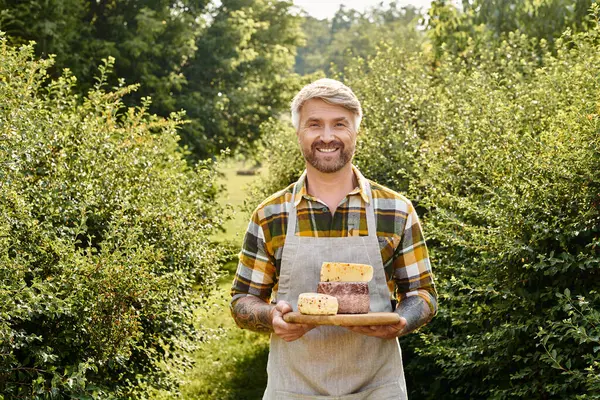 Alegre granjero guapo con tatuajes sosteniendo queso casero en sus manos y sonriendo a la cámara - foto de stock