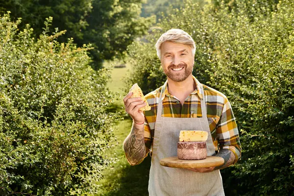 Cheerful handsome farmer with tattoos holding homemade cheese in his hands and smiling at camera — Stock Photo