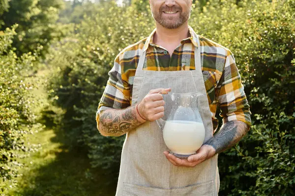 Vista ritagliata di agricoltore allegro con tatuaggi in possesso di vaso di latte fresco e sorridente felicemente — Foto stock
