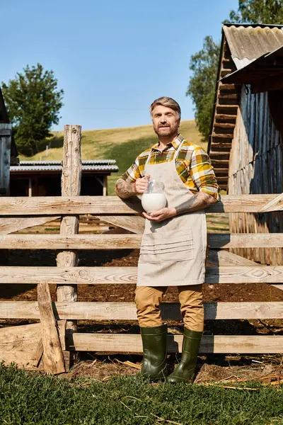 Hombre guapo con tatuajes en brazos sosteniendo frasco de leche fresca y mirando a la cámara mientras está en la granja - foto de stock