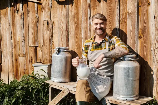 Cheerful attractive farmer sitting near his house with jar and churns of milk and smiling at camera — Stock Photo