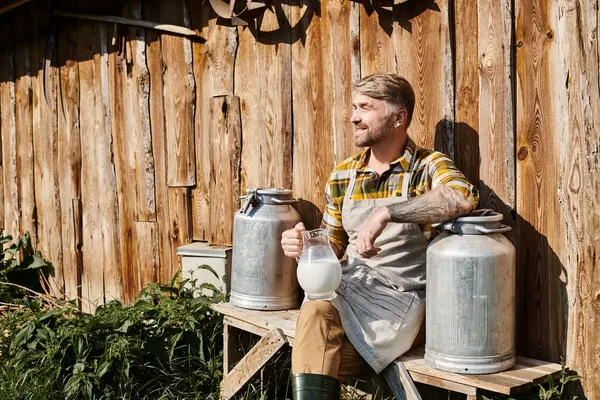 Cheerful attractive farmer sitting near his house with jar and churns of milk and smiling away — Stock Photo
