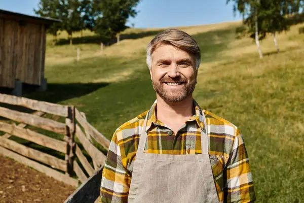 Guapo hombre alegre con tatuajes posando junto a la cerca y el estiércol en la granja y mirando a la cámara - foto de stock