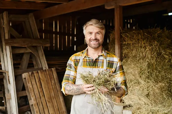 Alegre granjero bien parecido con barba y tatuajes sosteniendo un poco de heno y sonriendo a la cámara en la granja - foto de stock
