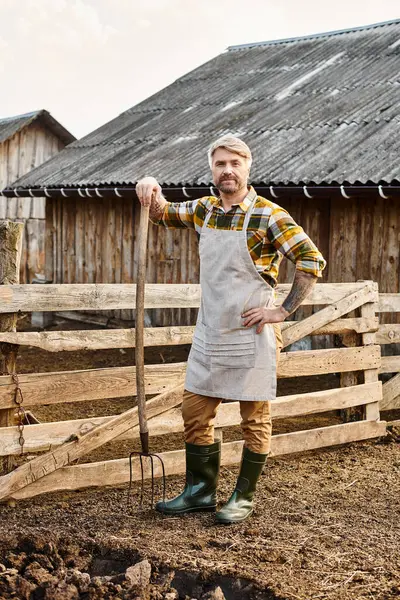 Bello agricoltore con tatuaggi utilizzando forcone mentre si lavora con letame e guardando la fotocamera — Foto stock