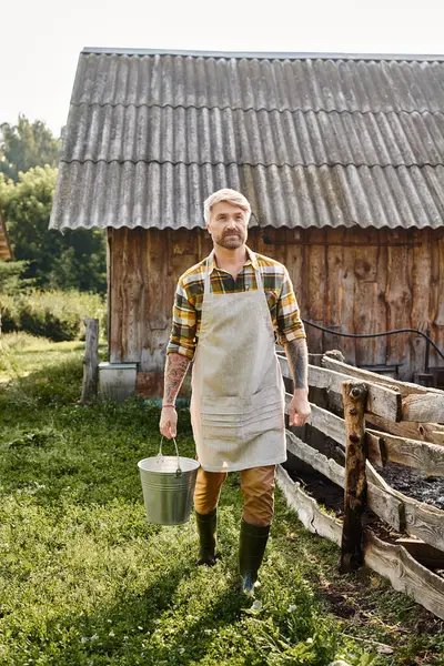 Hombre barbudo bien parecido con tatuajes en brazos sosteniendo cubo con leche fresca mientras está en su granja - foto de stock