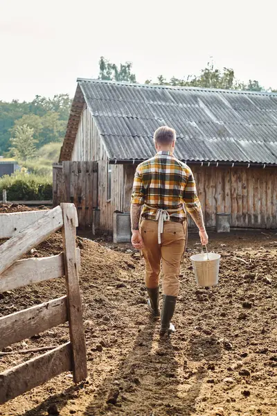 Vista posterior del hombre trabajador adulto con tatuajes en brazos sosteniendo cubo con leche mientras está en la granja - foto de stock