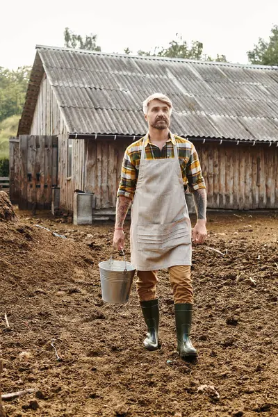 Hombre barbudo bien parecido con tatuajes en brazos sosteniendo cubo con leche fresca mientras está en su granja - foto de stock