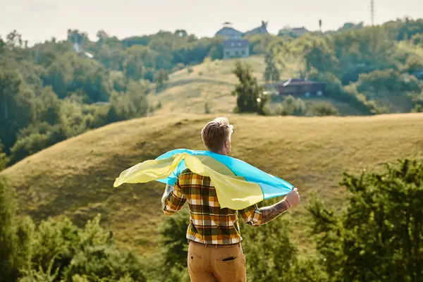 Back view of adult Ukrainian farmer with tattoos on arms posing with national flag, scenic landscape — Stock Photo