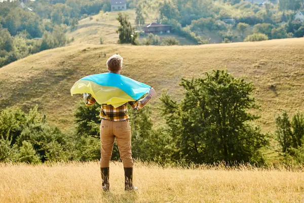 Back view of adult Ukrainian farmer with tattoos on arms posing with national flag, scenic landscape — Stock Photo