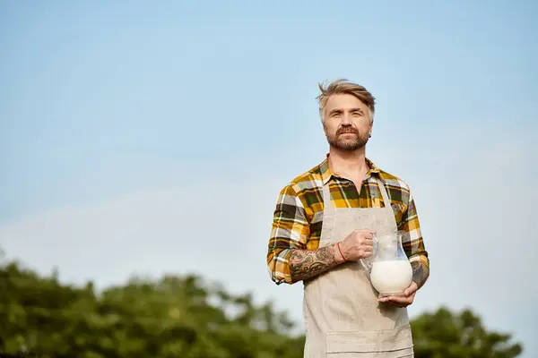 Granjero moderno de buen aspecto con barba y tatuajes sosteniendo frasco de leche fresca y mirando a la cámara - foto de stock
