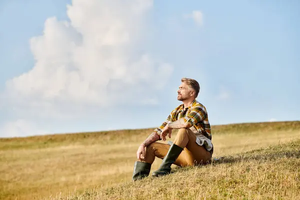 Hombre dedicado bien parecido con barba relajante en el campo de primavera verde y mirando hacia otro lado, agricultor moderno - foto de stock