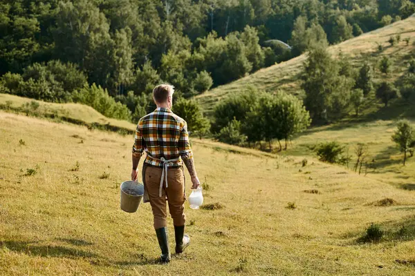 Vista trasera del trabajador agricultor moderno con tatuajes caminando con cubo y tarro de leche en las manos - foto de stock