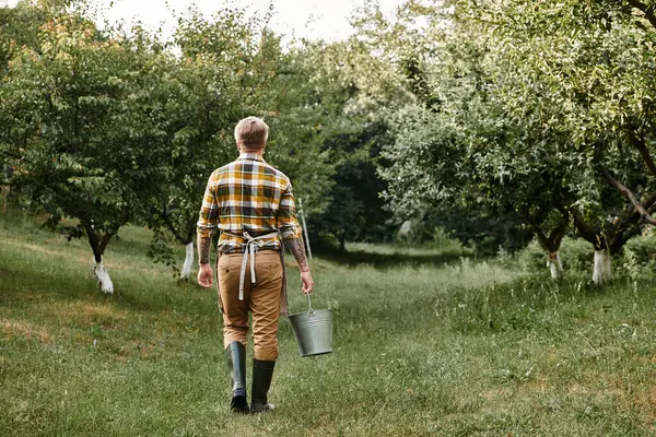 Back view homem trabalhador com tatuagens em braços trabalhando no jardim e segurando balde de metal na mão — Fotografia de Stock