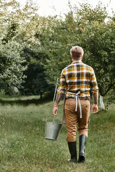 Vertical shot of hard working man with tattoos working in garden and holding metal bucket in hand — Stock Photo