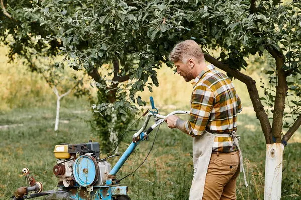 Handsome bearded man in casual attire with tattoos using lawnmower while in garden on his farm — Stock Photo