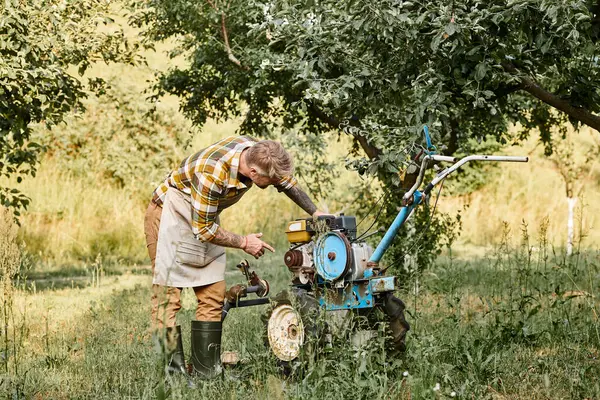 Bel homme barbu en tenue décontractée avec des tatouages en utilisant tondeuse à gazon tandis que dans le jardin sur sa ferme — Photo de stock