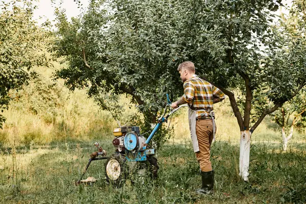 Homem barbudo bonito em traje casual com tatuagens usando cortador de grama enquanto no jardim em sua fazenda — Fotografia de Stock