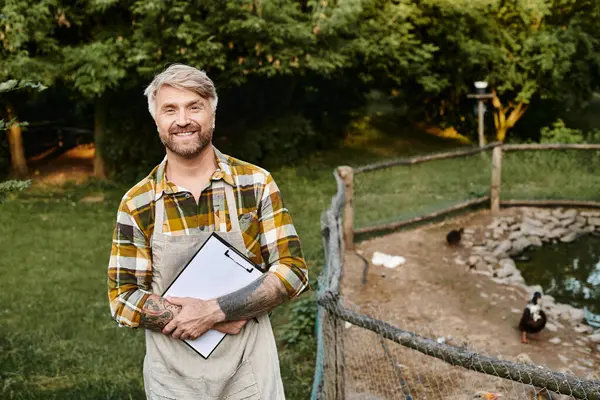 Guapo granjero alegre con tatuajes y barba sujetando portapapeles cerca de aviario y sonriendo a la cámara - foto de stock