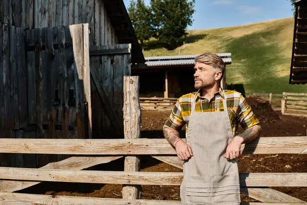 Привлекательный бородатый современный фермер в повседневной одежде с татуировками, позирующими и смотрящими в сторону в деревне — стоковое фото