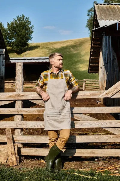Atractivo barbudo moderno agricultor en traje casual con tatuajes posando y mirando hacia otro lado en la aldea - foto de stock