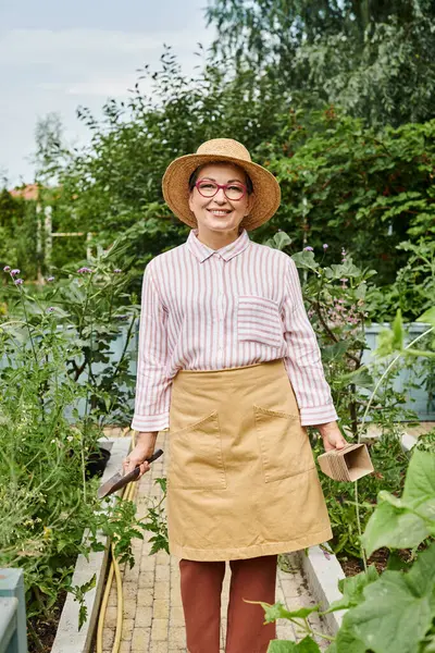 Hermosa mujer madura alegre en sombrero y atuendo casual sosteniendo herramientas de jardinería y sonriendo a la cámara - foto de stock