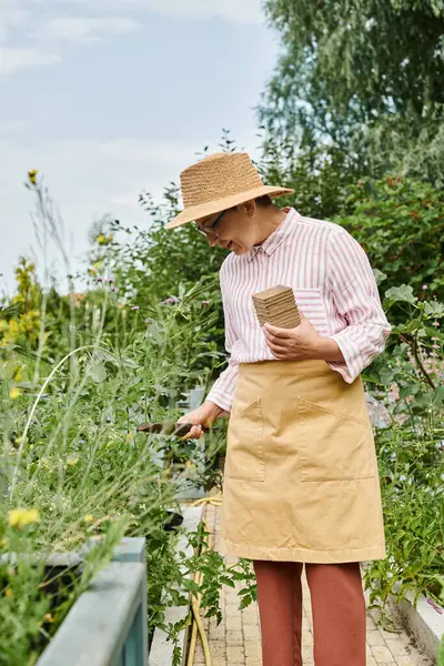 Atractiva mujer madura alegre con sombrero de paja usando sus herramientas de jardinería y sonriendo felizmente - foto de stock