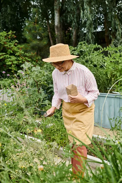 Приваблива весела зріла жінка з солом'яним капелюхом, використовуючи інструменти для садівництва та щасливо посміхаючись — стокове фото