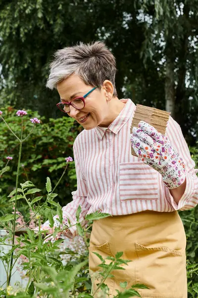 Atractiva mujer madura alegre con gafas y guantes usando equipo de jardinería en sus flores - foto de stock