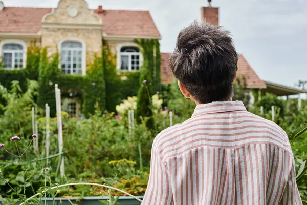 Vista trasera de la mujer madura con el pelo gris posando frente a su casa de pueblo moderno en Inglaterra - foto de stock
