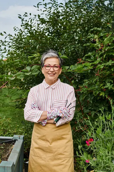 Jolly mature woman in casual attire with glasses holding rakes for gardening and smiling at camera — Stock Photo