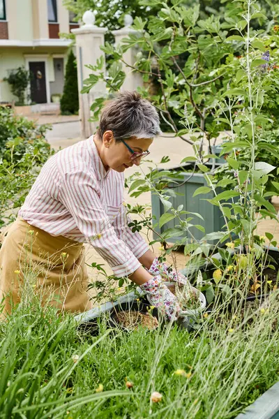 Attraktive, glückliche reife Frau mit Brille, die in ihrem lebhaften grünen Garten arbeitet und freudig lächelt — Stockfoto