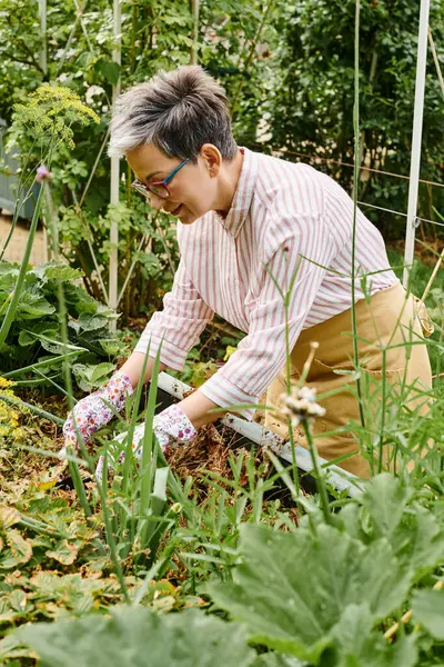 Belle femme mature heureuse avec des lunettes de travail dans son jardin vert vif et souriant joyeusement — Photo de stock