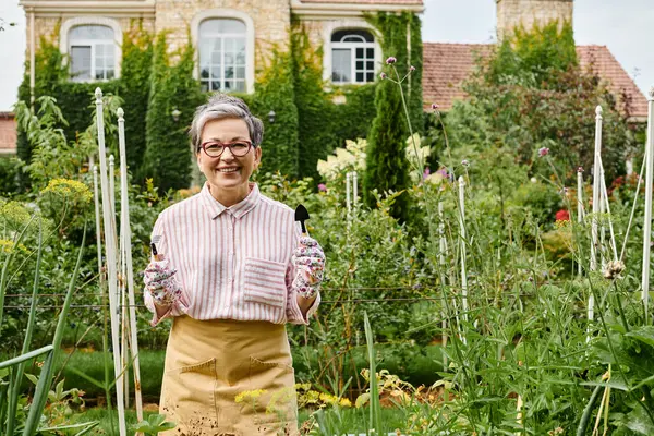 Cheerful mature woman with gloves and glasses holding gardening tools in hands and smiling at camera — Stock Photo